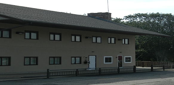 A two-story beige building with multiple windows, two main doors, and a fenced area in front, photographed during daylight.